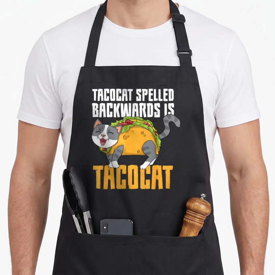 Tacocat mexican apron - Cartoon gray and white cat wearing a taco costume with palindrome text above and below — shown on a black cotton canvas apron against a white background