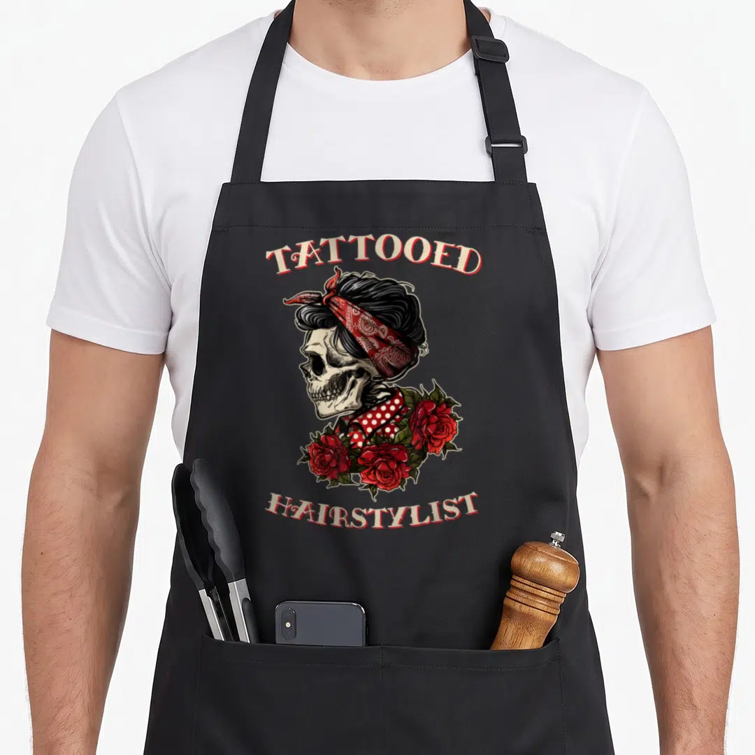 Skull hairstylist apron - Cartoon skull in red bandana with roses and arched tattooed hairstylist text — shown on a black cotton canvas apron against a white background