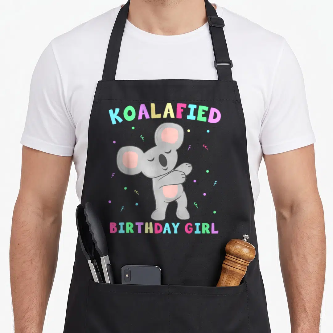 Koalafied bear apron - Cartoon grey koala dabbing with colorful confetti and arched text reading KOALAFIED BIRTHDAY GIRL — shown on a black cotton canvas apron against a white background