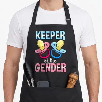 Keeper pacifier apron - Two cartoon pacifiers in pink blue yellow white with KEEPER of the GENDER text — shown on a black cotton canvas apron against a white background