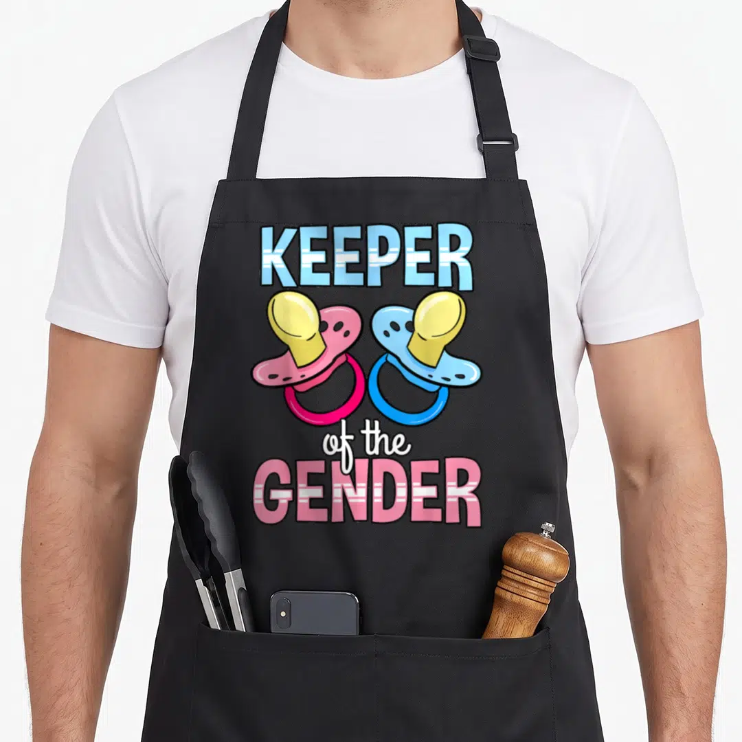 Keeper pacifier apron - Two cartoon pacifiers in pink blue yellow white with KEEPER of the GENDER text — shown on a black cotton canvas apron against a white background