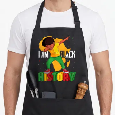 Funny month history apron - Cartoon boy dabbing on 3D HISTORY text in front of orange Africa map with white I AM BLACK — shown on a black cotton canvas apron against a white background