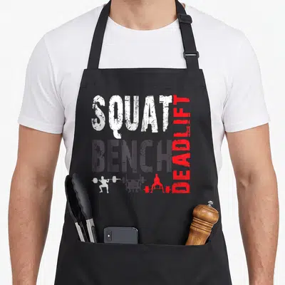 Powerlifting bench apron - SQUAT BENCH DEADLIFT text in white grey red with three weightlifter silhouettes performing — shown on a black cotton canvas apron against a white background