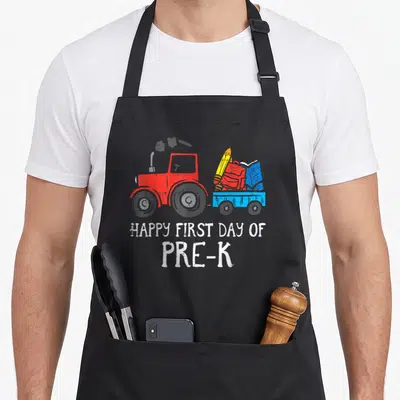 Tractor truck apron - Red tractor pulls blue trailer with backpack pencil and book under white text — shown on a black cotton canvas apron against a white background
