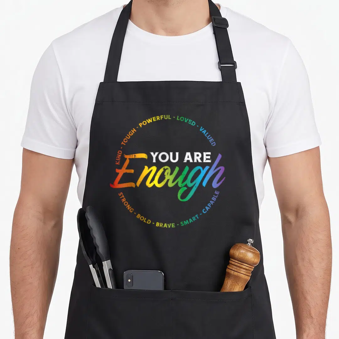Enough rainbow apron - Rainbow gradient script Enough text surrounded by circular positive affirmations in block letters — shown on a black cotton canvas apron against a white background
