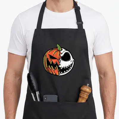 Face pumpkin apron - Circular cartoon graphic split between orange jack-o-lantern and white skeleton skull with black — shown on a black cotton canvas apron against a white background
