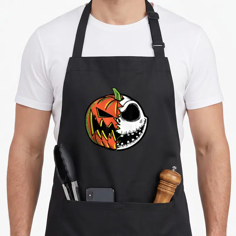 Face pumpkin apron - Circular cartoon graphic split between orange jack-o-lantern and white skeleton skull with black — shown on a black cotton canvas apron against a white background