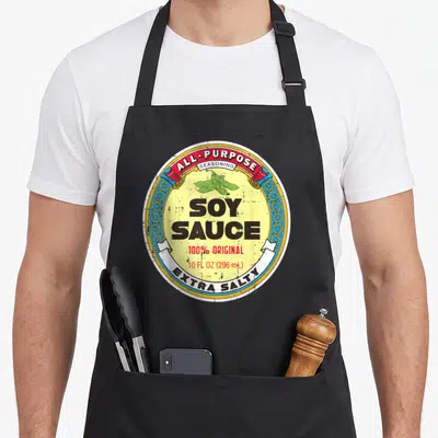 Soy halloween apron - Retro circular soy sauce label with soybeans, shakers, and text all-purpose seasoning extra salty — shown on a black cotton canvas apron against a white background