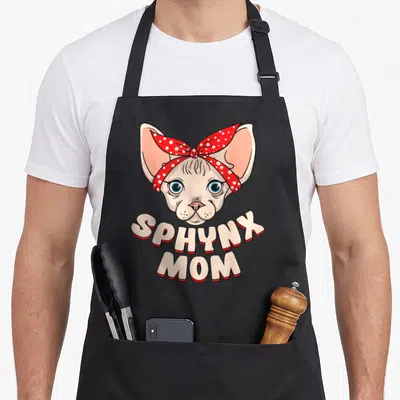 Mother cat apron - Cartoon Sphynx cat head with red polka dot bandana and beige text reading SPHYNX MOM — shown on a black cotton canvas apron against a white background