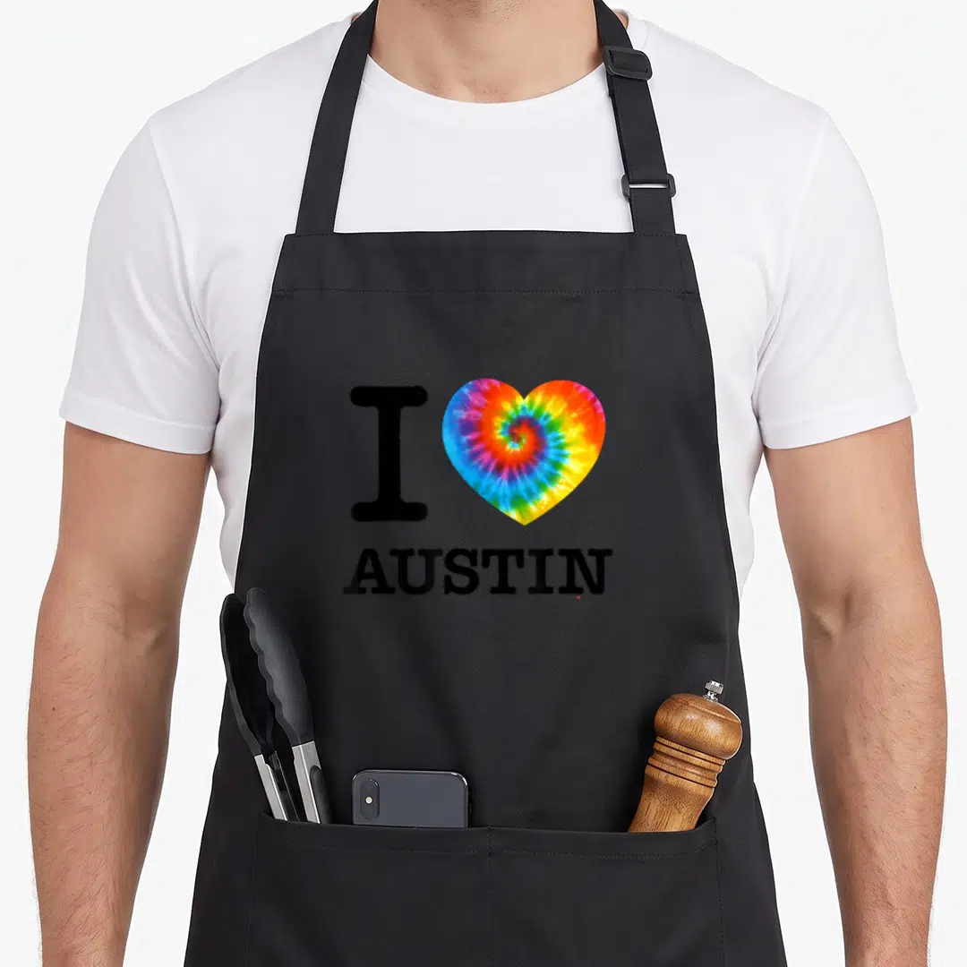 Austin tie-dye apron - Black I and AUSTIN text flanking a multicolor tie-dye heart — shown on a black cotton canvas apron against a white background