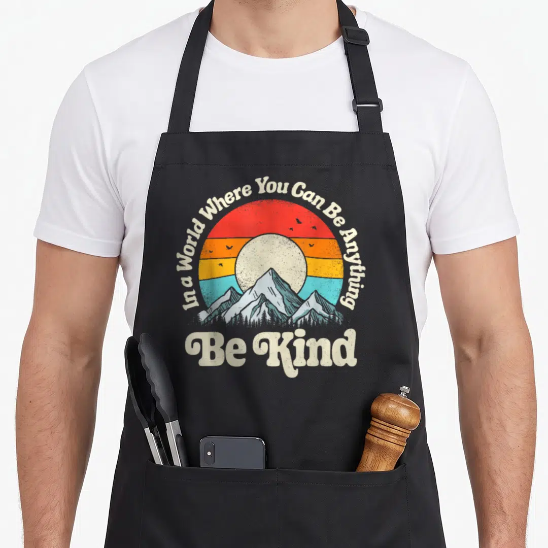 Kind novelty apron - Retro circular sunset with mountains birds and text In a World Where You Can Be Anything Be Kind — shown on a black cotton canvas apron against a white background