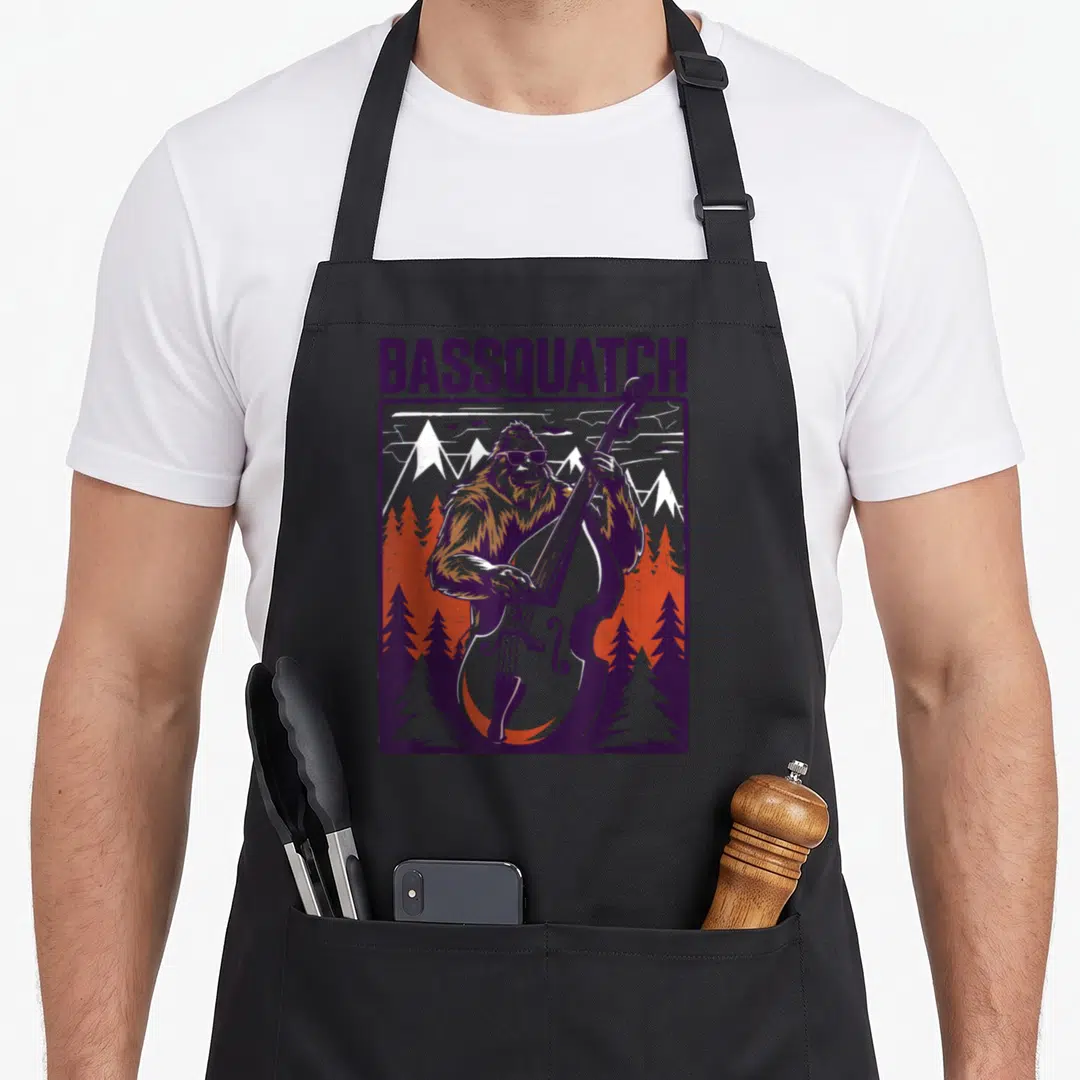 Double bigfoot apron - Bassquatch text above cartoon Sasquatch playing double bass in forest scene — shown on a black cotton canvas apron against a white background