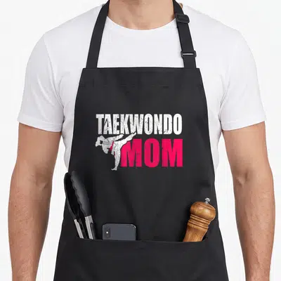 Cute taekwondo apron - Distressed white TAEKWONDO text above pink MOM with a kicking martial artist silhouette — shown on a black cotton canvas apron against a white background