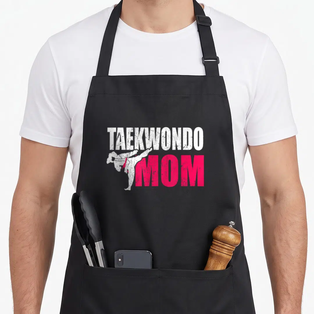 Cute taekwondo apron - Distressed white TAEKWONDO text above pink MOM with a kicking martial artist silhouette — shown on a black cotton canvas apron against a white background