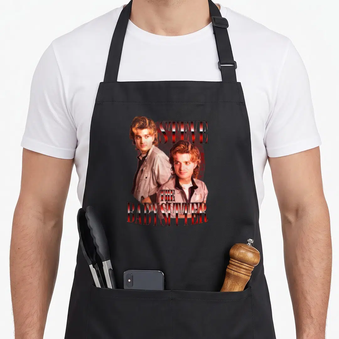 Baby stranger apron - Retro print of two Steve Harrington portraits with red STEVE THE BABYSITTER text on grey — shown on a black cotton canvas apron against a white background
