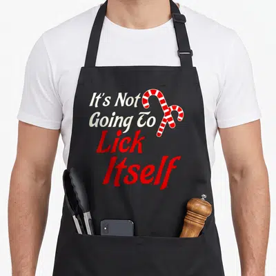 Lick christmas apron - It's Not Going To Lick Itself text with red and white candy cane graphic — shown on a black cotton canvas apron against a white background