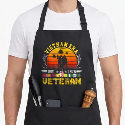 Veteran retired soldier apron - Distressed Vietnam Era 1961 1975 veteran text with soldier silhouettes and ribbon bar — shown on a black cotton canvas apron against a white background