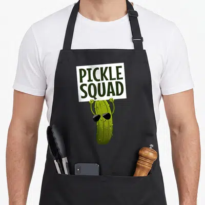 Retro cucumber pickle apron - Cartoon green pickle wearing sunglasses holding a white sign reading PICKLE SQUAD — shown on a black cotton canvas apron against a white background