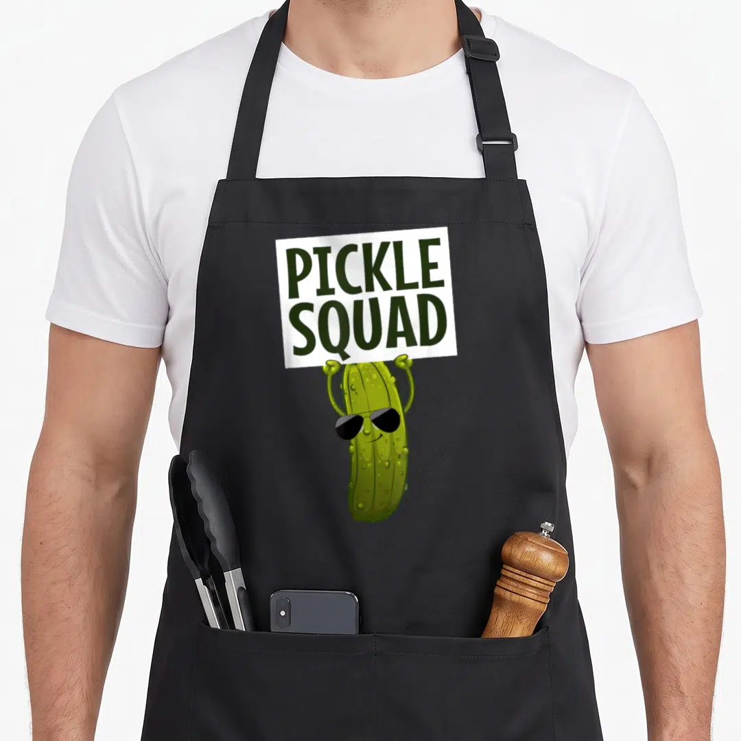 Retro cucumber pickle apron - Cartoon green pickle wearing sunglasses holding a white sign reading PICKLE SQUAD — shown on a black cotton canvas apron against a white background