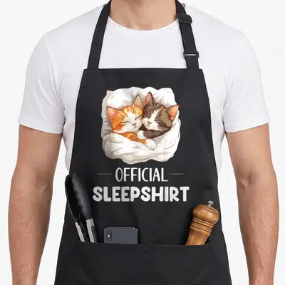 Sleepshirt cat apron - Two curled orange and grey cats in white blanket with official sleepshirt text — shown on a black cotton canvas apron against a white background