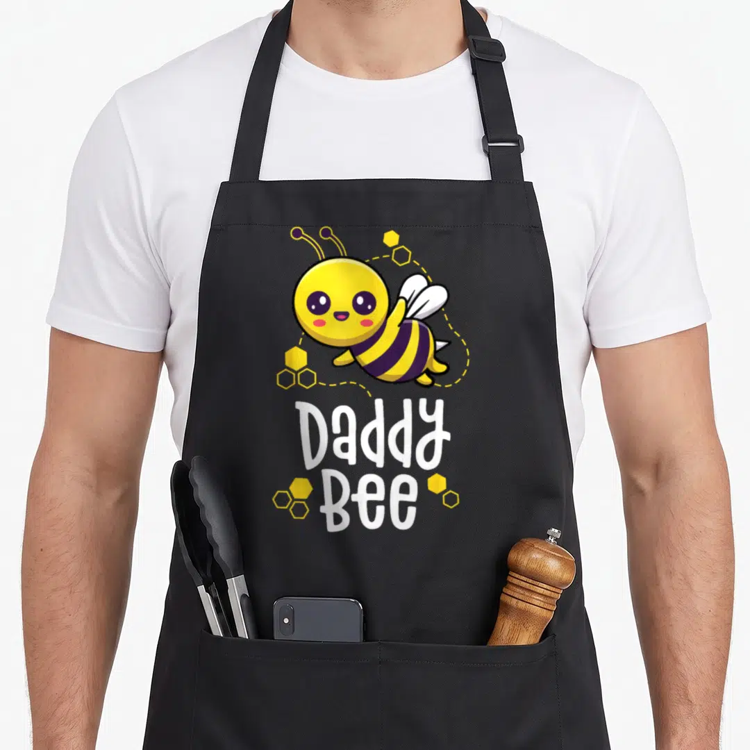 Family daddy bee aprons - Cartoon bee with yellow hexagons and white Daddy Bee text — shown on a black cotton canvas apron against a white background