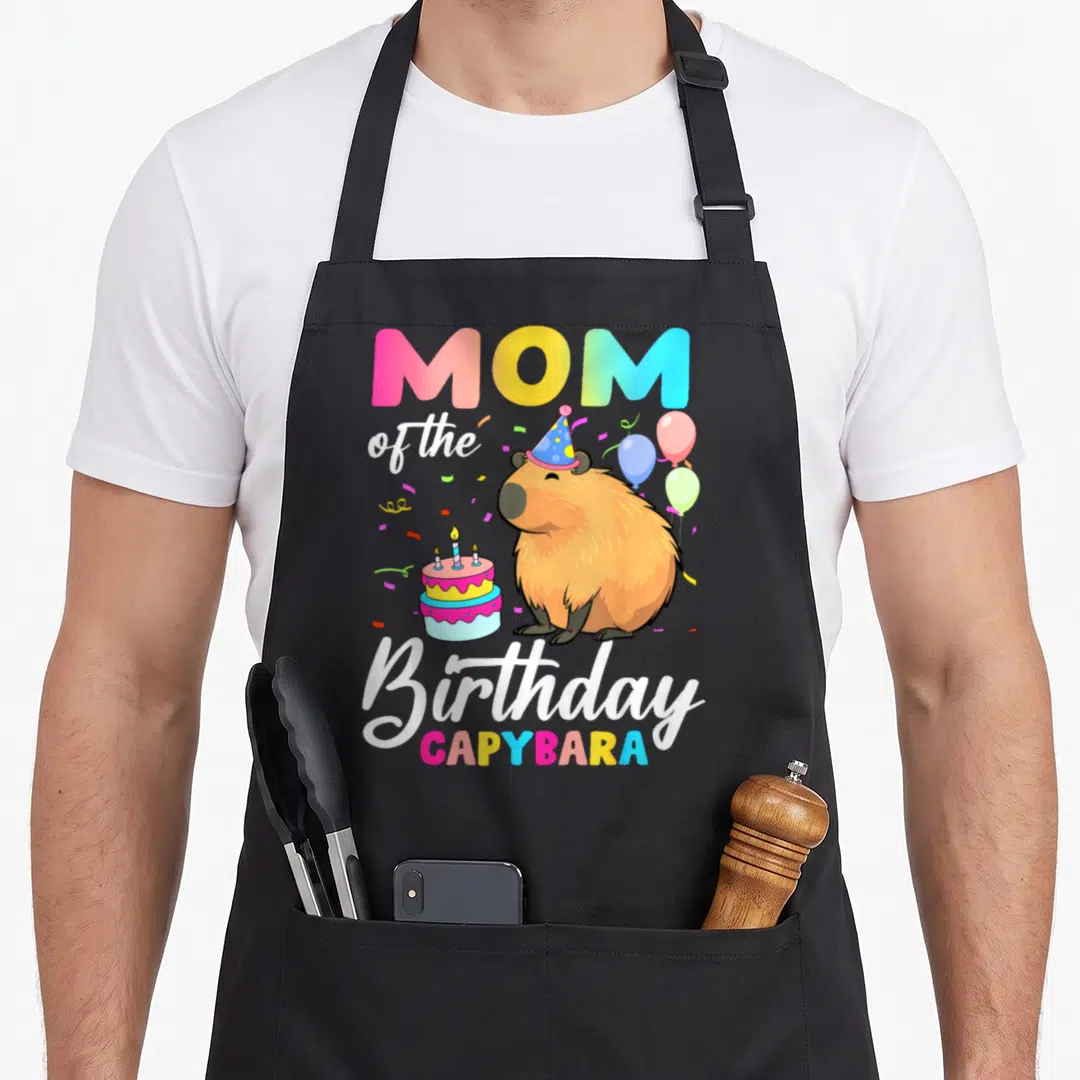 Capybara family aprons - Mom of the Birthday Capybara text with capybara wearing party hat near cake and balloons — shown on a black cotton canvas apron against a white background