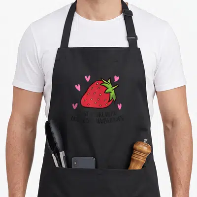 Cute strawberry apron - Red cartoon strawberry with green top and four pink hearts above black text reading JUST A GIRL — shown on a black cotton canvas apron against a white background