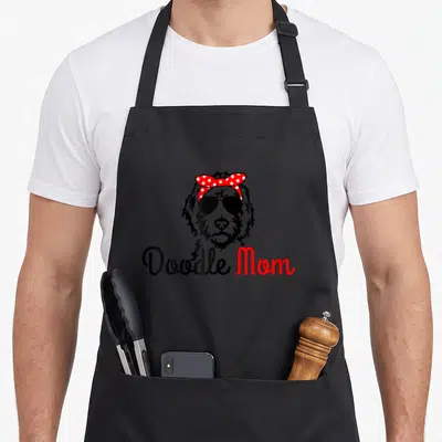 Doodle goldendoodle dog apron - Line art doodle dog wearing sunglasses and red polka dot bow above Doodle Mom text — shown on a black cotton canvas apron against a white background