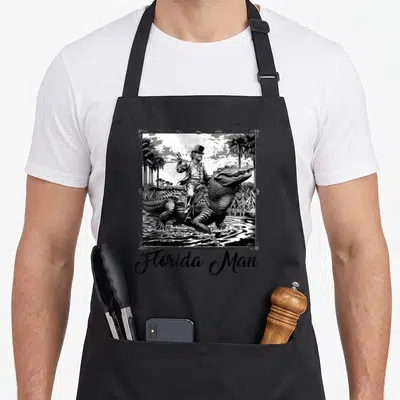 Riding florida apron - Man in top hat riding alligator through water with palm trees and Florida Man text — shown on a black cotton canvas apron against a white background