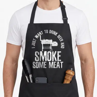 Drink beer meat aprons - White distressed text and smoker grill graphic on black fabric — shown on a black cotton canvas apron against a white background