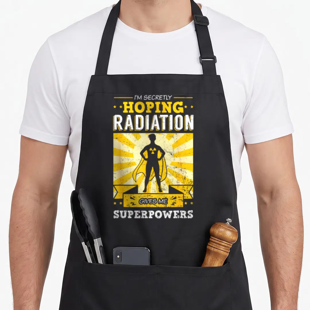 Hoping radiation aprons - Grey superhero silhouette on yellow sunburst with hope radiation superpowers text — shown on a black cotton canvas apron against a white background