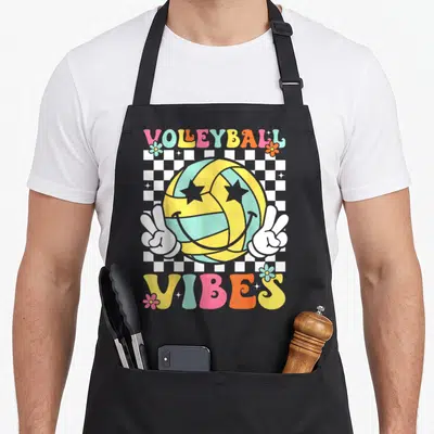 Funny volleyball game aprons - VOLEYBALL VIBES text above a smiling volleyball with peace hands on a checkerboard — shown on a black cotton canvas apron against a white background