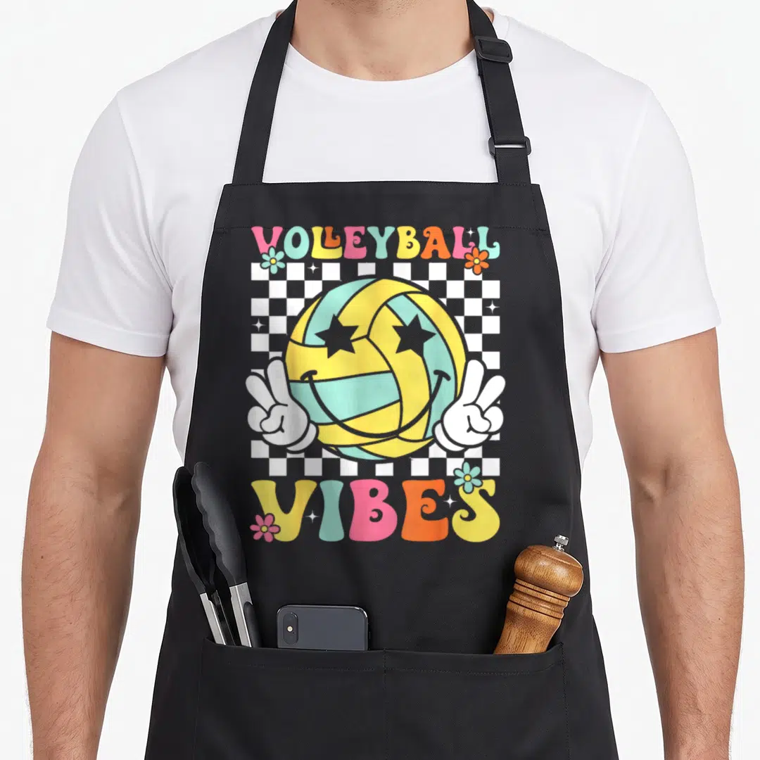 Funny volleyball game aprons - VOLEYBALL VIBES text above a smiling volleyball with peace hands on a checkerboard — shown on a black cotton canvas apron against a white background