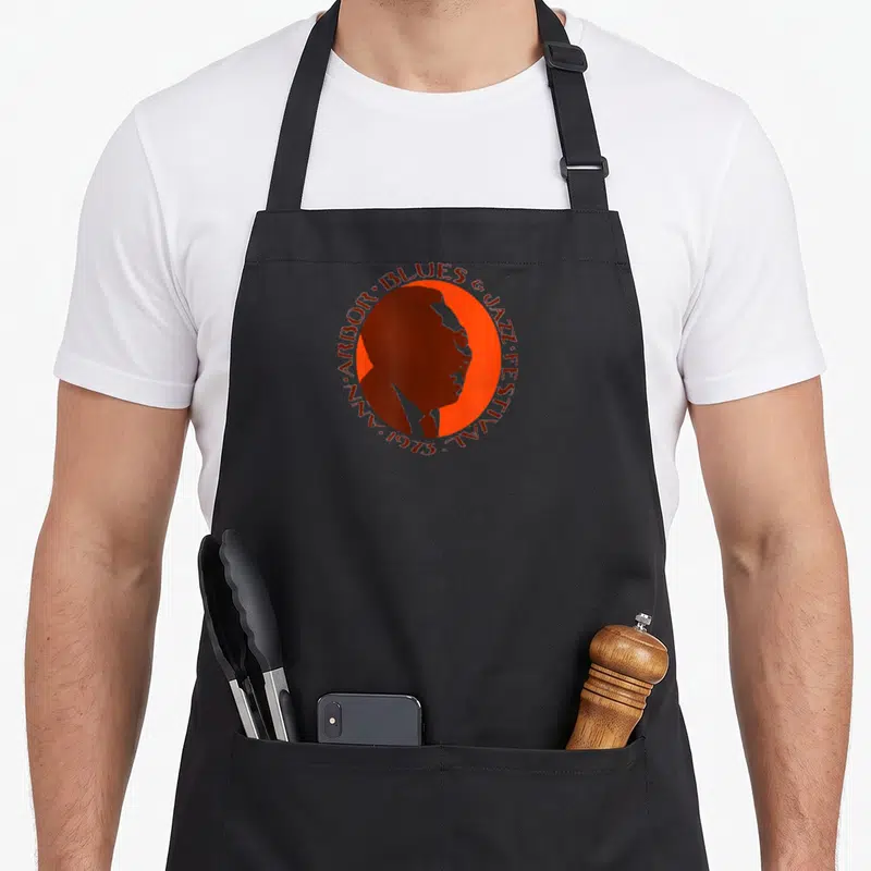 Ann jazz apron - Brown man profile inside orange circle with 1973 Ann Arbor Blues & Jazz Festival text border — shown on a black cotton canvas apron against a white background