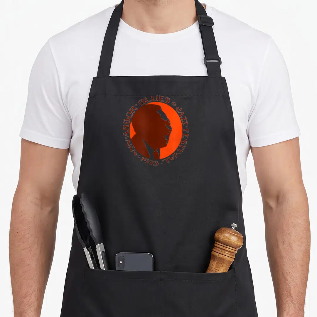 Ann jazz apron - Brown man profile inside orange circle with 1973 Ann Arbor Blues & Jazz Festival text border — shown on a black cotton canvas apron against a white background