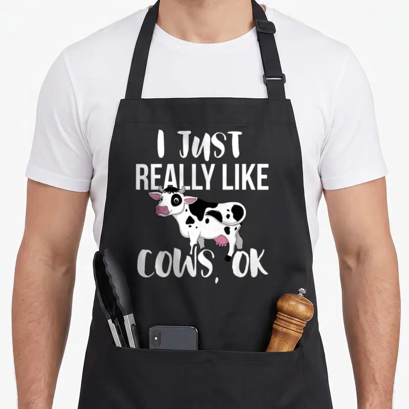 Cow rancher farmer apron - Cartoon cow with I Just Really Like Cows OK text in white black and pink — shown on a black cotton canvas apron against a white background