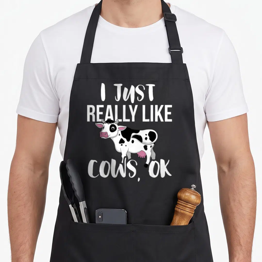 Cow rancher farmer apron - Cartoon cow with I Just Really Like Cows OK text in white black and pink — shown on a black cotton canvas apron against a white background