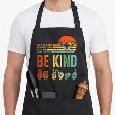 Anything autism apron - Retro sun butterfly puzzle pieces six sign language hands be kind text — shown on a black cotton canvas apron against a white background