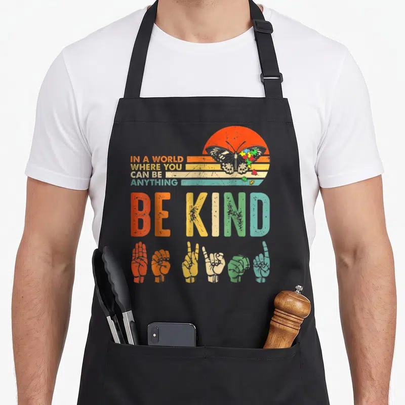Anything autism apron - Retro sun butterfly puzzle pieces six sign language hands be kind text — shown on a black cotton canvas apron against a white background
