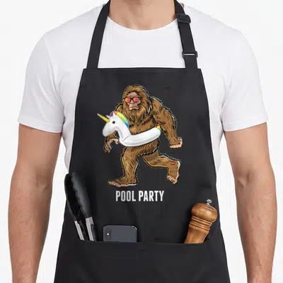 Pool bigfoot apron - Cartoon brown Bigfoot with sunglasses holding white unicorn floatie and POOL PARTY text — shown on a black cotton canvas apron against a white background