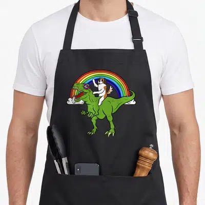 Rex dog apron - Cartoon saint bernard riding green t-rex with purple sunglasses under a rainbow — shown on a black cotton canvas apron against a white background