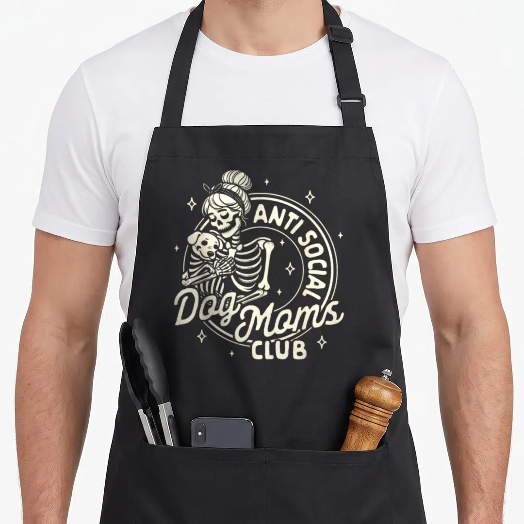 Skeleton dog apron - Beige line art of skeleton woman hugging skeleton dog with anti social dog moms club text — shown on a black cotton canvas apron against a white background