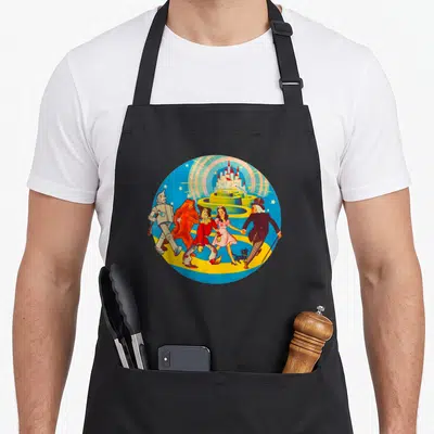 Lion dorothy apron - Cartoon Wizard of Oz characters walking on yellow path toward castle in circular blue frame — shown on a black cotton canvas apron against a white background