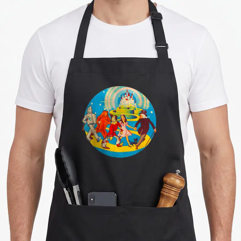 Lion dorothy apron - Cartoon Wizard of Oz characters walking on yellow path toward castle in circular blue frame — shown on a black cotton canvas apron against a white background