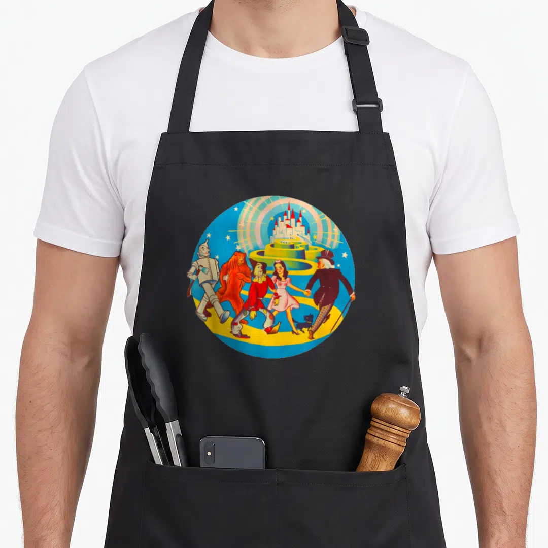 Lion dorothy apron - Cartoon Wizard of Oz characters walking on yellow path toward castle in circular blue frame — shown on a black cotton canvas apron against a white background