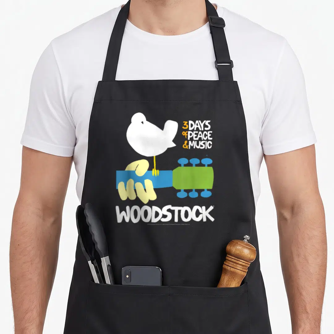 Woodstock peace apron - White bird on blue green guitar neck with 3 days of peace and music text — shown on a black cotton canvas apron against a white background
