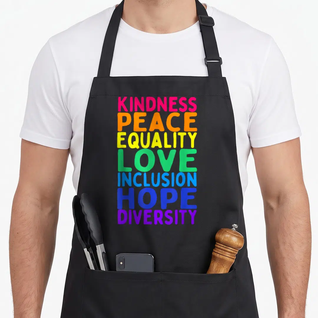 Kindness celebrate diversity apron - Stacked rainbow text reading kindness peace equality love inclusion hope diversity — shown on a black cotton canvas apron against a white background
