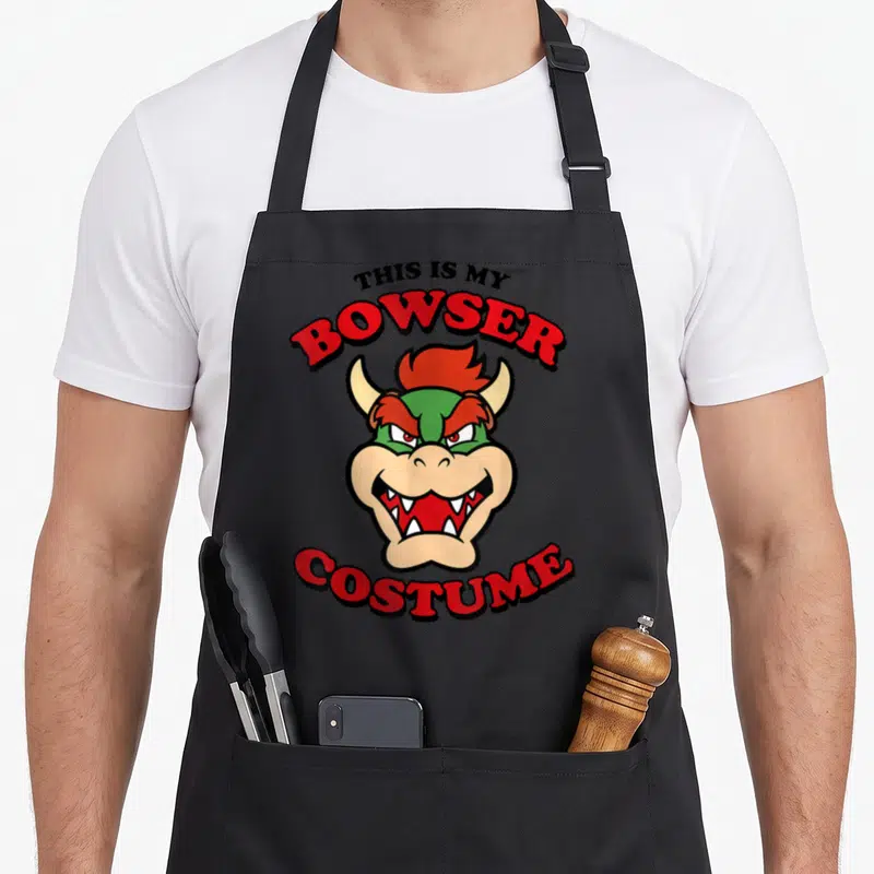 Costume game over thirty apron - Cartoon Bowser head with red and green text reading This Is My Bowser Costume arched — shown on a black cotton canvas apron against a white background