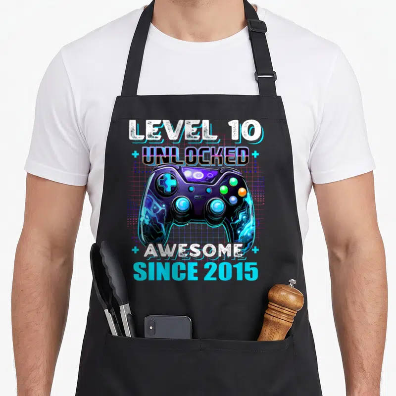 10th birthday level video game apron - Retro game controller with LEVEL 10 UNLOCKED AWESOME SINCE 2015 text in purple — shown on a black cotton canvas apron against a white background