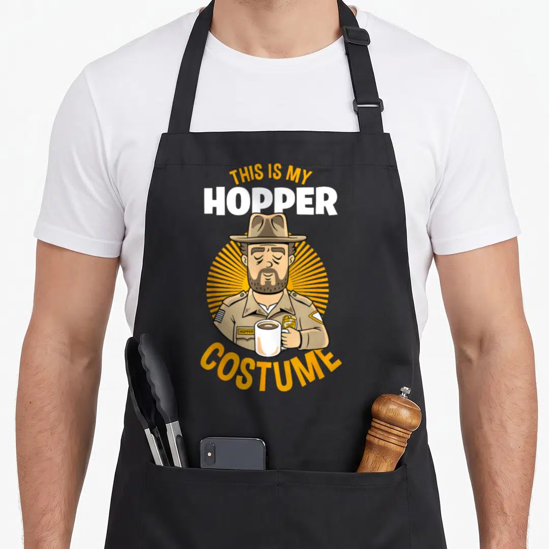 Hopper halloween apron - Cartoon man in brown uniform holding white mug with text THIS IS MY HOPPER COSTUME — shown on a black cotton canvas apron against a white background